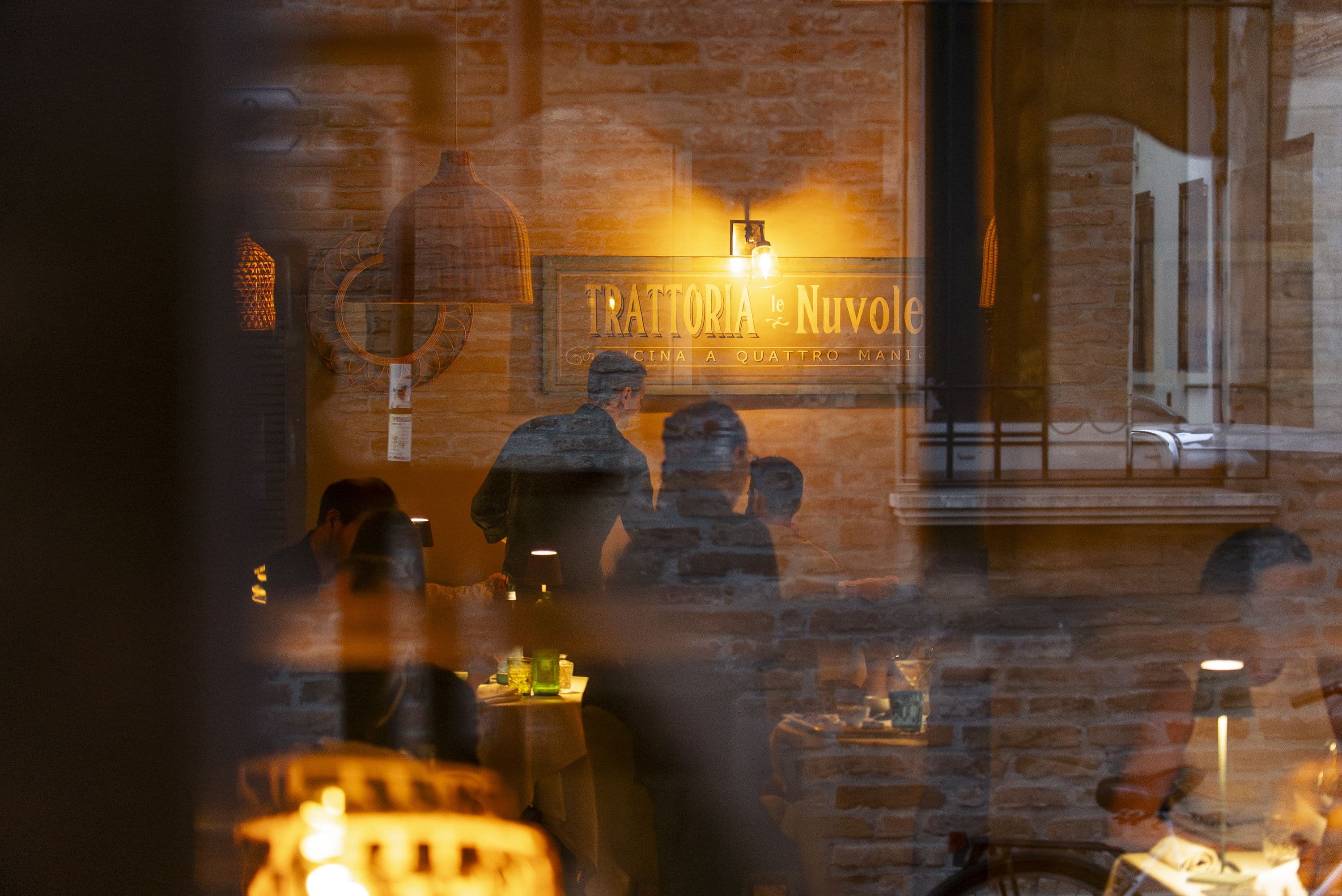 The dining room of Trattoria Le Nuvole seen from the street in the evening, with the sign reading «Cucina a quattro mani»